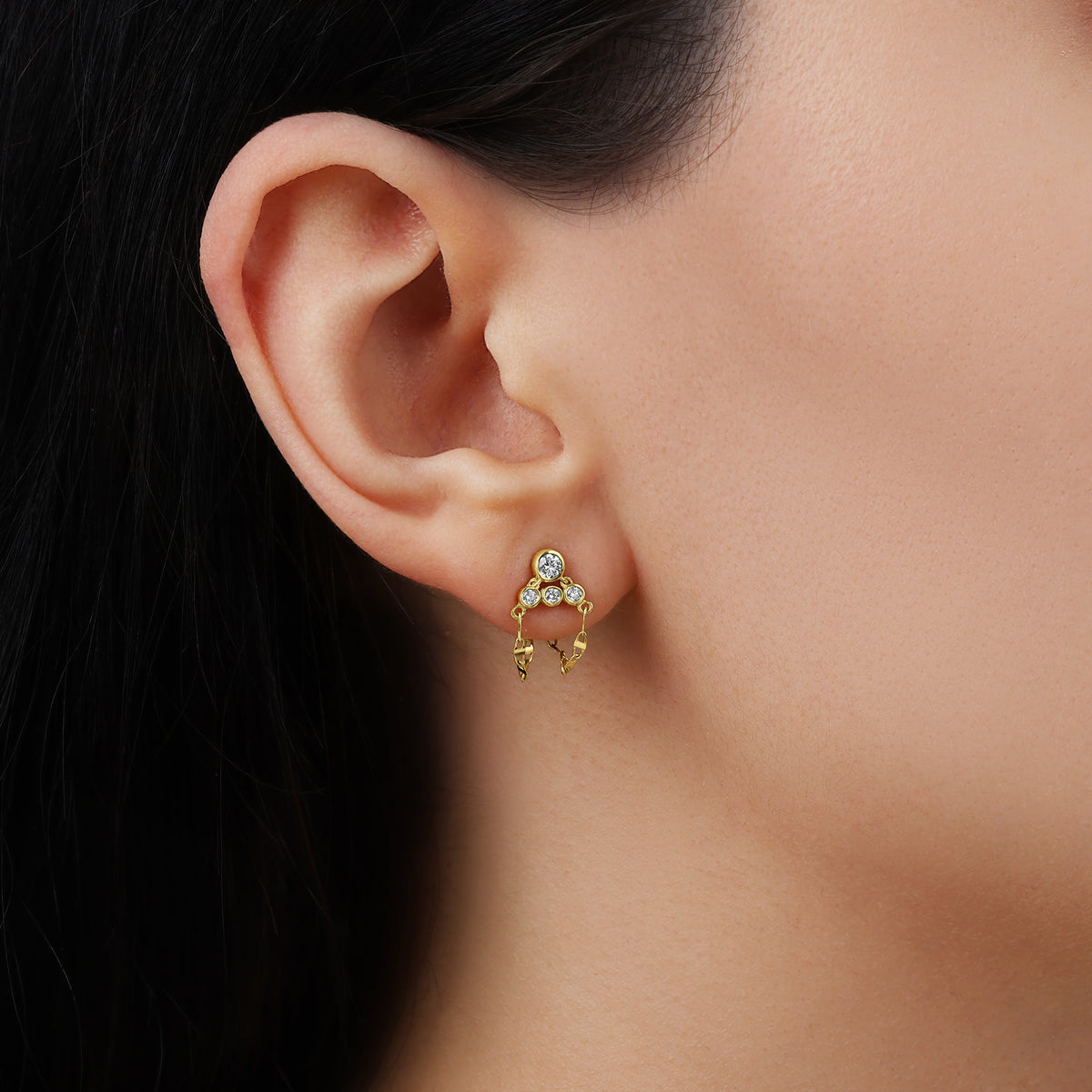 A close-up side view of a model wearing a fine 14k yellow gold post drop earring. The earring features a tiered cluster of four round bezel-set white diamond gemstones with three delicate loops of mariner link chain dangling gracefully from the earlobe.
