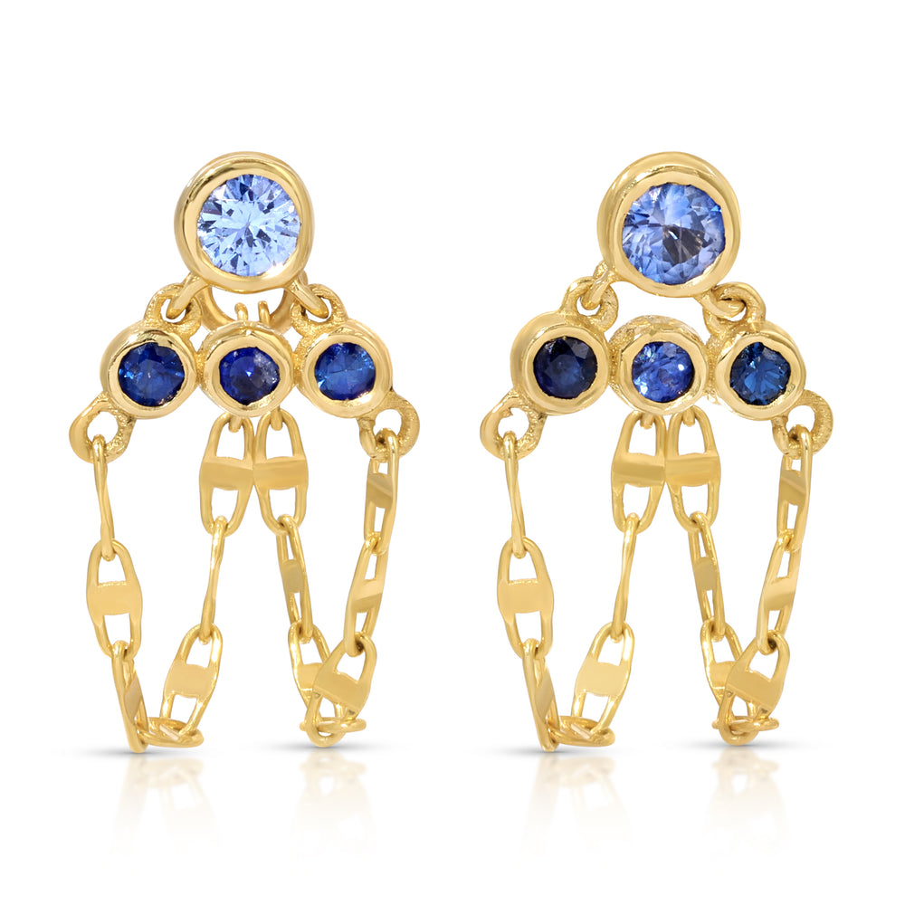 A pair of handcrafted 14k yellow gold Anna drop earrings featuring a tiered geometric setting with light blue and dark blue natural sapphires, suspended above delicate looping strands of mariner link chain against a plain white background.