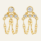 Pair of drop earrings featuring four round-cut brilliant white diamond gemstones bezel-set in polished 14 karat yellow gold, arranged with one top stone leading down to a triangular cluster of three stones with two dangling marine link chains that wrap from the front under the earlobe to connect to the back post of the pushback closure, shown on a solid uniform light cream background.
