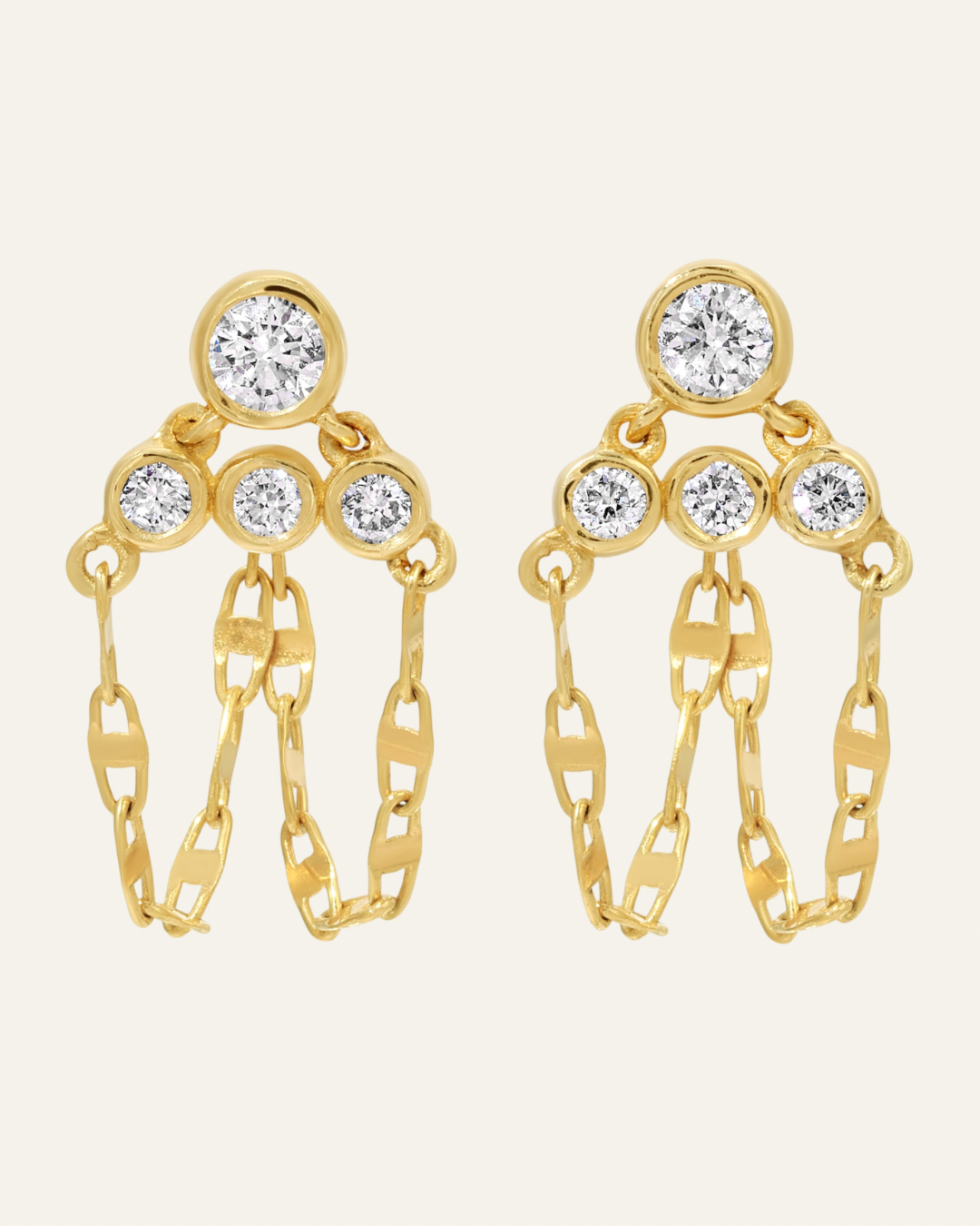 Pair of drop earrings featuring four round-cut brilliant white diamond gemstones bezel-set in polished 14 karat yellow gold, arranged with one top stone leading down to a triangular cluster of three stones with two dangling marine link chains that wrap from the front under the earlobe to connect to the back post of the pushback closure, shown on a solid uniform light cream background.